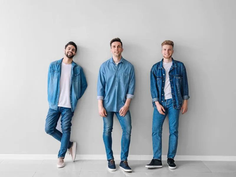 Três homens de jeans e jaqueta encostados em parede clara